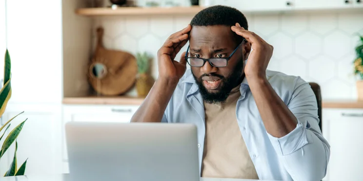 A man with a horrified expression looks at a laptop, while sitting in the kitchen