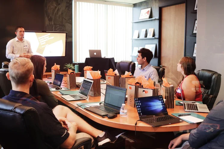 five individuals sitting around a table with lots of laptops on it, listening to someone present slides during a project planning meeting