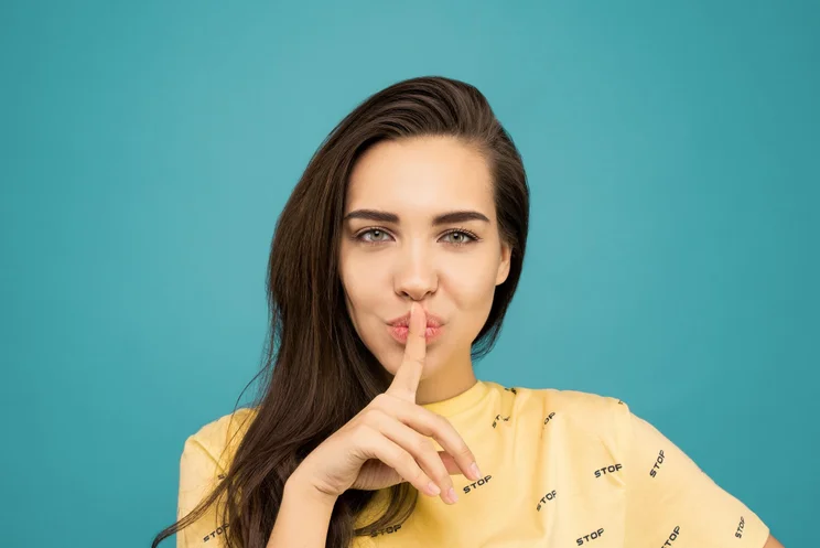 Woman in a yellow top on a turquoise background with her finger on her lips to signify quiet