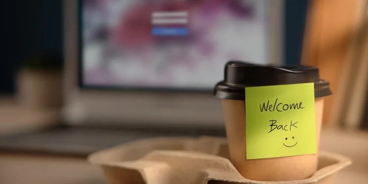 Coffee cup with note saying "Welcome Back" on desk illustrating return to office concept