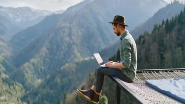 Young man in hat working remotely whilst travelling in beautiful mountain area
