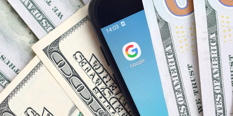 Phone with Google icon nestled in middle of US dollar bills illustrating Google and money