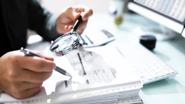 Man with magnifying glass and pen going over paperwork illustrative of asset management compliance.