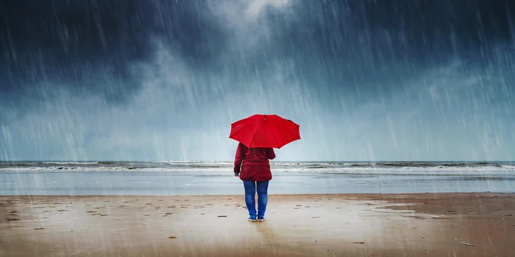 Person standing with an umbrella in the rain on a beach.