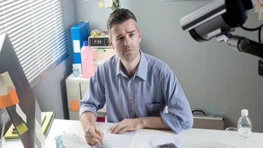 A man working remotely at his desk being spied on by his employer and CCTV