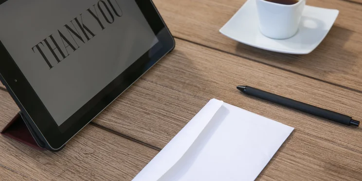 An ipad with the phrase thank you on it, next to a cup of coffee, pen, and envelope.
