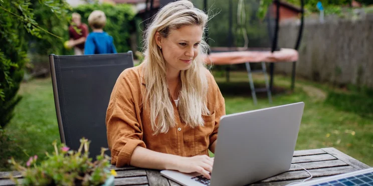 Woman remote working in her backyard with her two kids playing in the background.