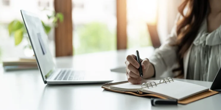 A woman sits at her desk, watching an online course and taking notes.