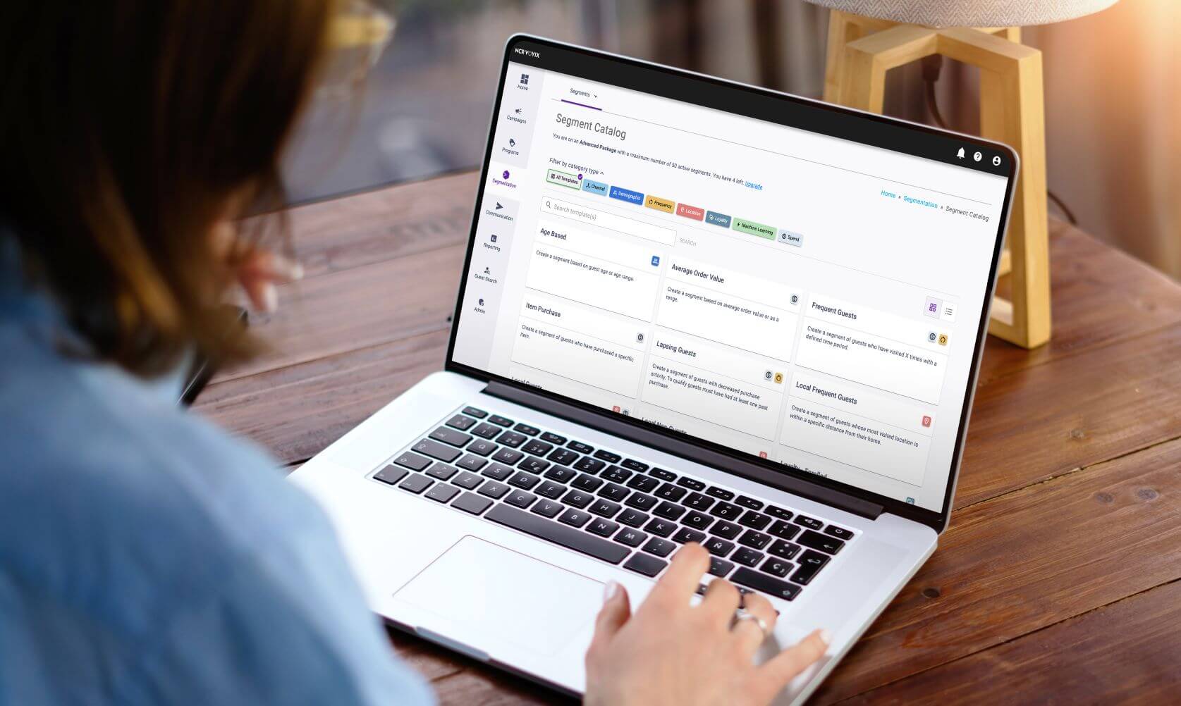 An over-the-shoulder view of a person using a laptop on a wooden desk. The screen shows the Aloha Consumer Marketing "Segment Catalog" dashboard, featuring customer segmentation options, filter tags, and detailed target audience cards.
