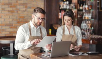 Two restaurant managers wearing beige aprons sitting at a wooden table in a bar setting, reviewing paperwork together alongside an open laptop and a tablet.