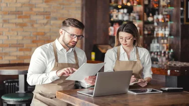 Two restaurant managers wearing beige aprons sitting at a wooden table in a bar setting, reviewing paperwork together alongside an open laptop and a tablet.