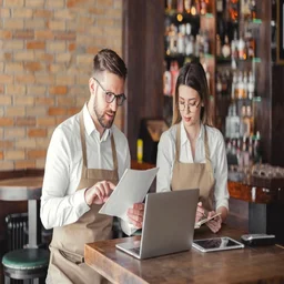 Two restaurant managers wearing beige aprons sitting at a wooden table in a bar setting, reviewing paperwork together alongside an open laptop and a tablet.