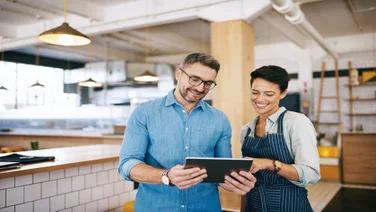 A male business owner in a blue shirt holding a tablet alongside a female employee in a striped apron, both looking down at the tablet screen and smiling inside a modern restaurant.
