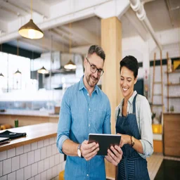 A male business owner in a blue shirt holding a tablet alongside a female employee in a striped apron, both looking down at the tablet screen and smiling inside a modern restaurant.