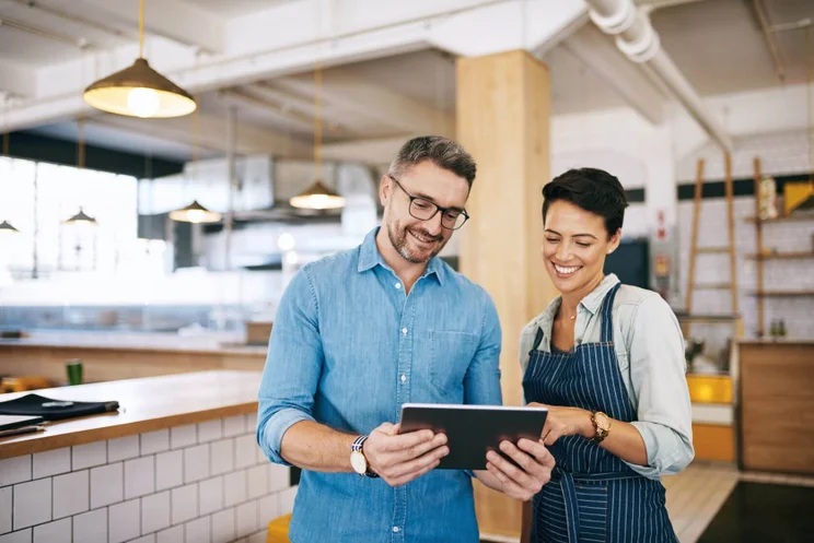 A male business owner in a blue shirt holding a tablet alongside a female employee in a striped apron, both looking down at the tablet screen and smiling inside a modern restaurant.
